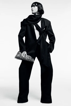 Fashion editorial photo of a woman posing in a black blazer and trousers, holding a beaded clutch in a white photo studio.