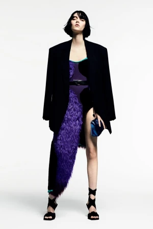 Model with bob haircut wears oversized black blazer, purple textured skirt, and black lace-up heels against a plain white background.