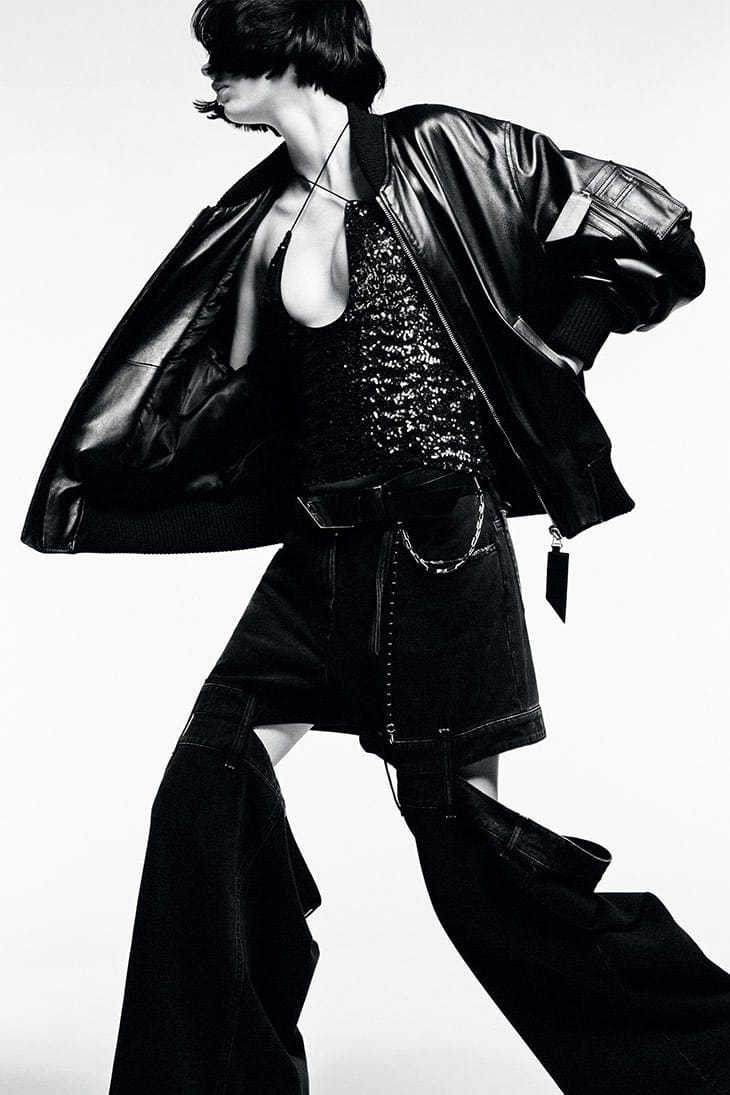 Fashion editorial shot of a woman in a black leather jacket, sequined top, and deconstructed denim pants against a white background.