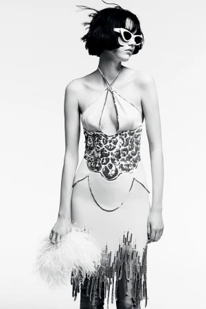 A woman in an elegant halter dress with sequin detailing poses against a white backdrop, holding a feathered accessory, in black and white.