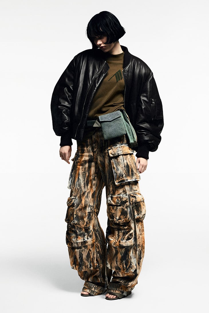 Woman in an olive t-shirt and black bomber jacket poses in a photo studio wearing brown and black patterned cargo pants.