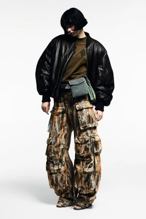 Woman in an olive t-shirt and black bomber jacket poses in a photo studio wearing brown and black patterned cargo pants.