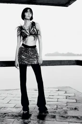 Model Steinberg poses in a feathered bra top, sequined mini skirt, and black flared trousers outdoors in black and white.