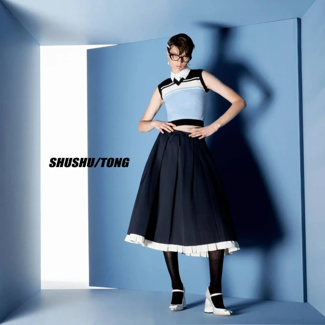 Fashion model poses in a blue-themed studio, wearing a cropped sweater vest, pleated skirt, tights, and heels for a Shushu/Tong shoot.