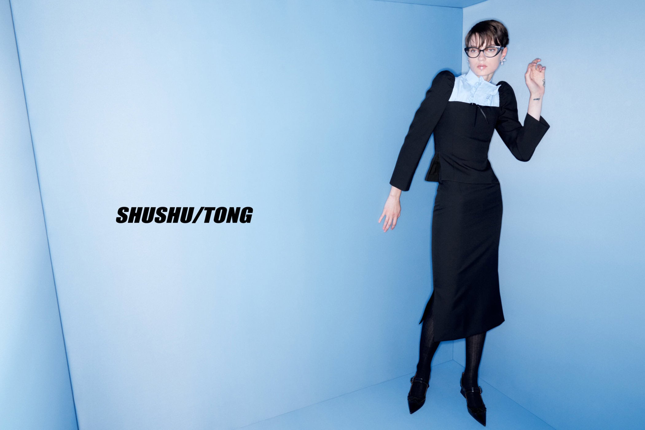 Fashion model in a black skirt suit and eyewear poses in a blue corner photo studio, capturing a modern and stylish editorial look.