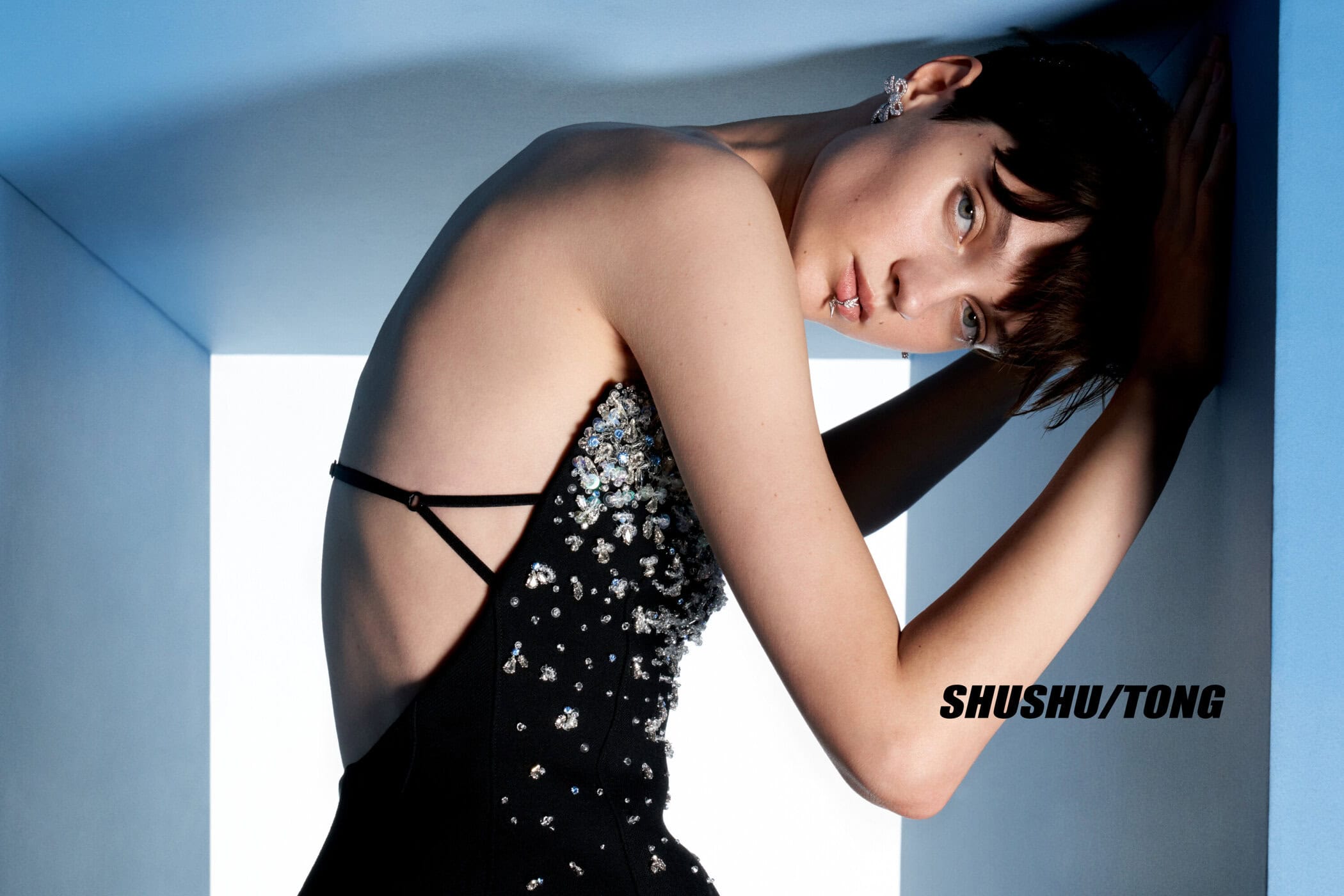A woman with dark hair is dressed in a black dress with jeweled accents, leaning against a blue wall. Fashion photography, studio setting.