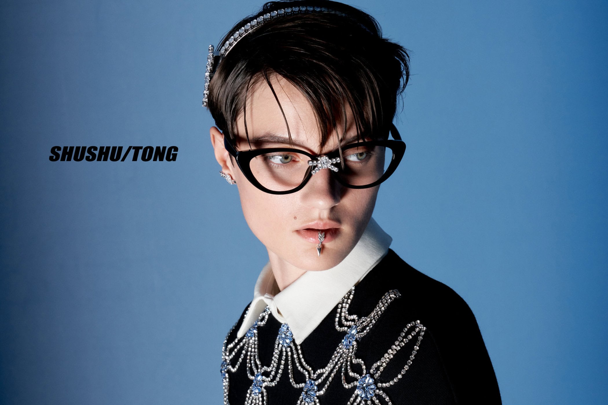 Portrait of a woman with short hair, wearing glasses, jewelry, and a black and white top, set against a blue background.