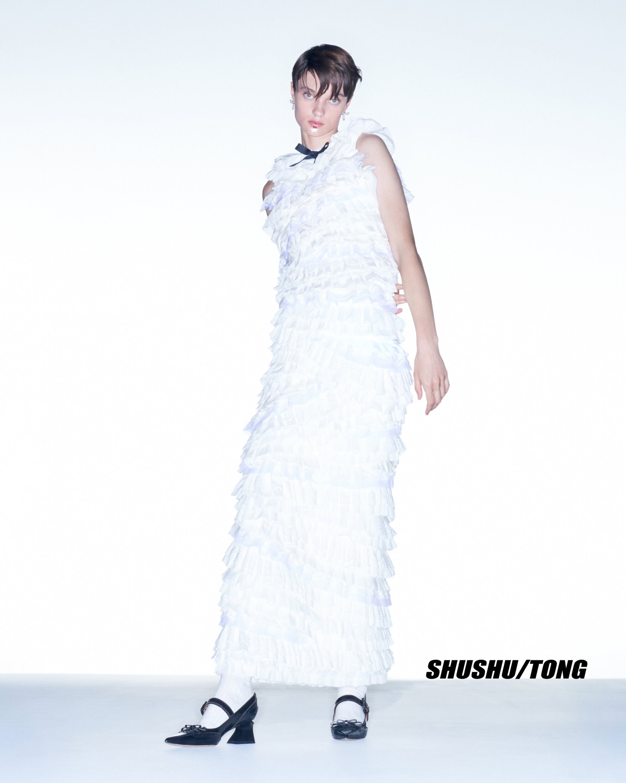Full-length studio portrait of a slender, androgynous person in a long, tiered, ruffled, white dress and black Mary Janes with white socks.