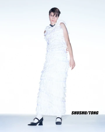 Full-length studio portrait of a slender, androgynous person in a long, tiered, ruffled, white dress and black Mary Janes with white socks.