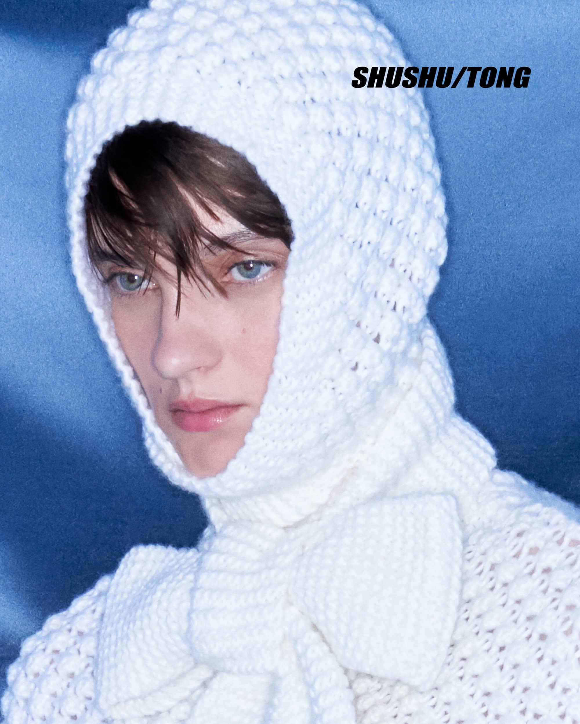 Close-up of a model wearing a white crocheted hood and scarf with bow detail against a soft blue background in a fashion setting.