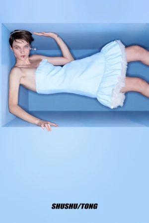 Woman in a pale blue dress with white ruffles lies in a blue box, studio shot, fashion, pale skin, androgynous, SHUSHU/TONG branding.
