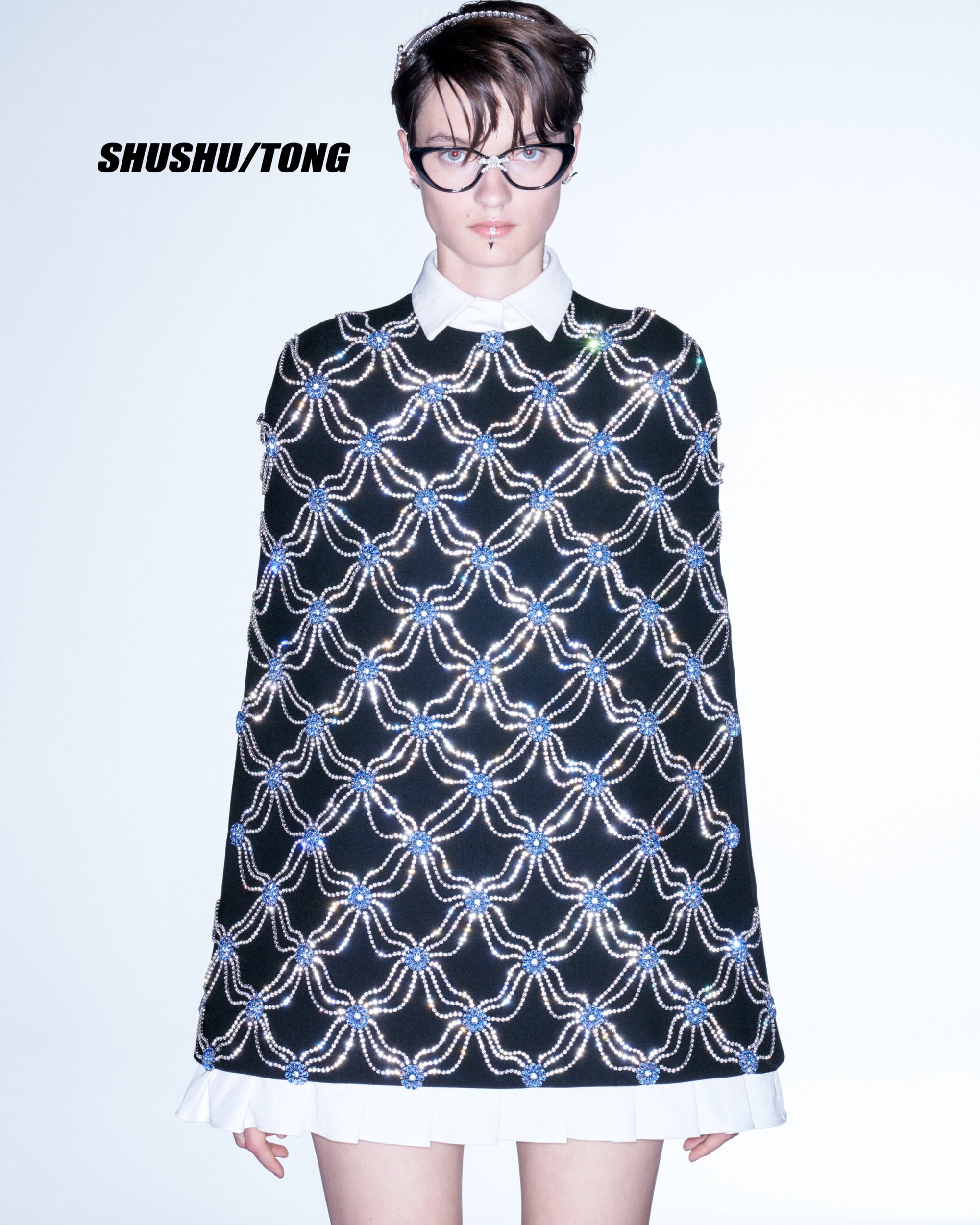 Woman wears a black dress with blue floral pattern and a white collared shirt. The dress has a cape-like design and extends to mid-thigh.