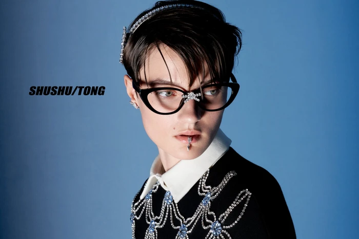 Model Tanya Churbanova wearing crystal-embellished cat-eye glasses, lip jewelry, and a beaded black knit top against a blue background.