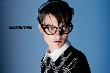 Model Tanya Churbanova wearing crystal-embellished cat-eye glasses, lip jewelry, and a beaded black knit top against a blue background.