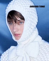 Model Tanya Churbanova wearing a textured white knit balaclava hood tied with a bow against a blue backdrop.