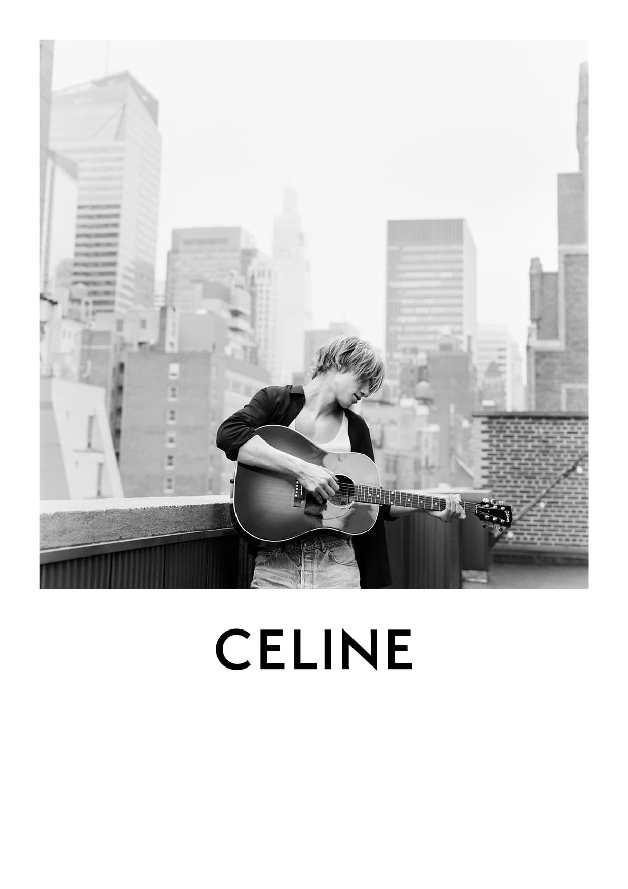 Black-and-white portrait of a young adult playing the guitar on a rooftop with city buildings in the background and the brand CELINE.