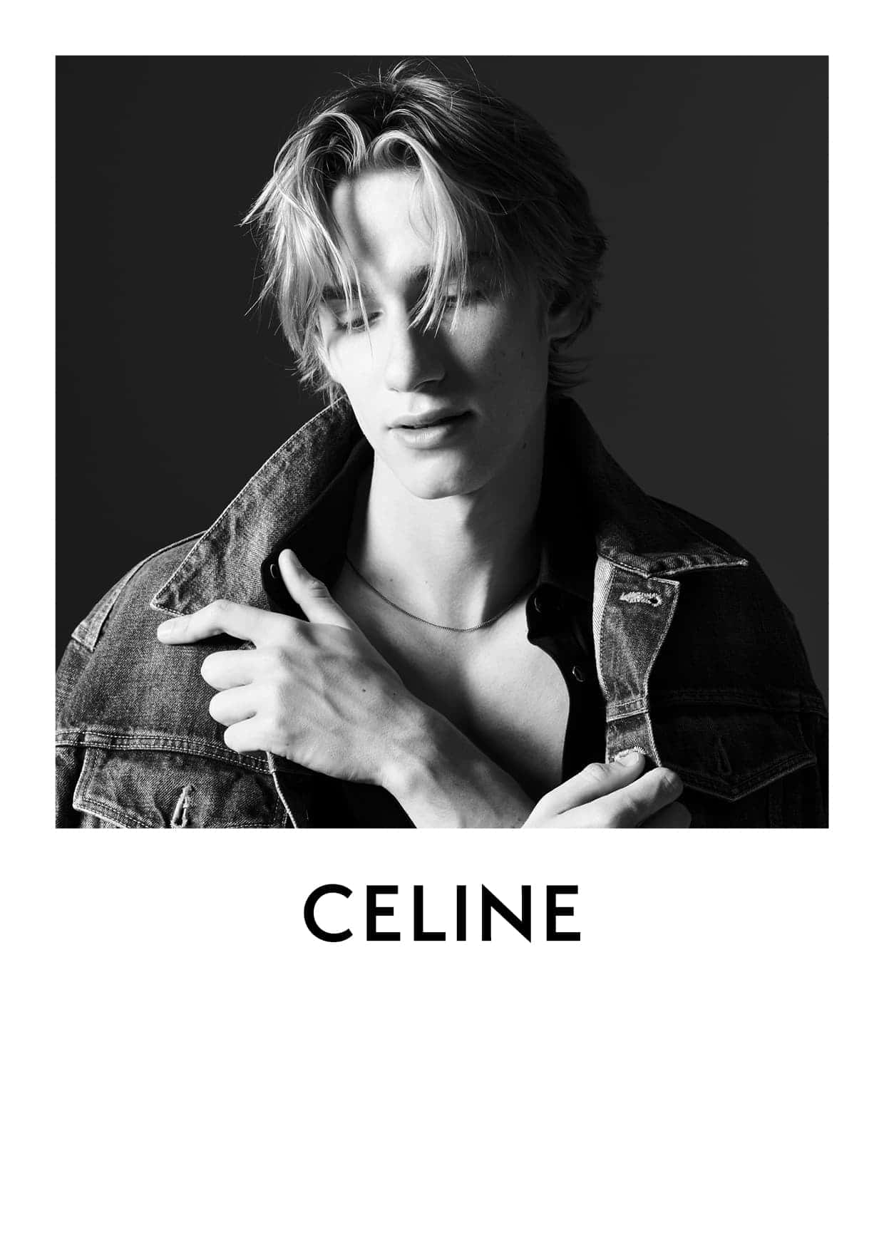 Monochrome portrait of a young adult male model with blond hair wearing a denim jacket looking downward in a studio setting.