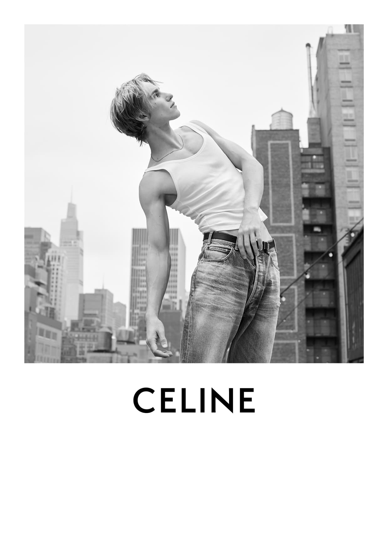 A male fashion model in a white tank top and blue jeans poses on a rooftop with a cityscape background for a black-and-white fashion editorial.