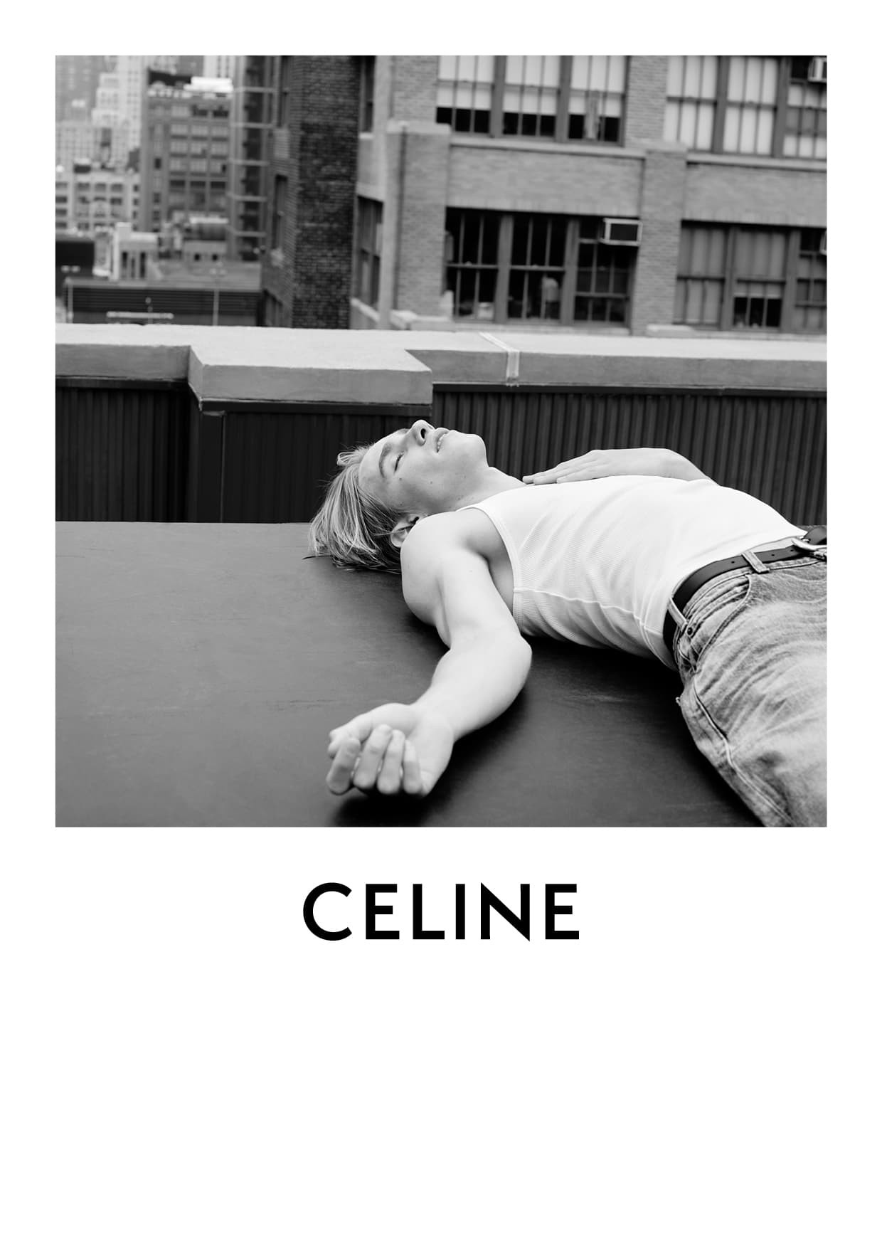 Black and white outdoor fashion shot of a young adult male laying down wearing jeans and a tank top with city buildings in the background.
