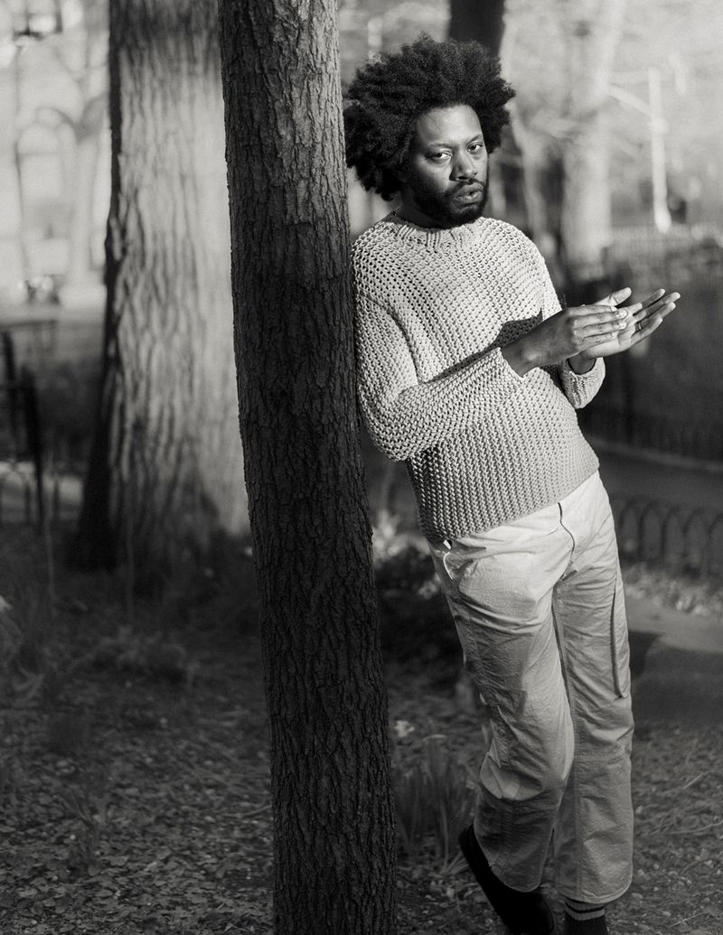Black and white portrait of a Black adult male leaning against a tree trunk in an outdoor setting, wearing a sweater and pants.