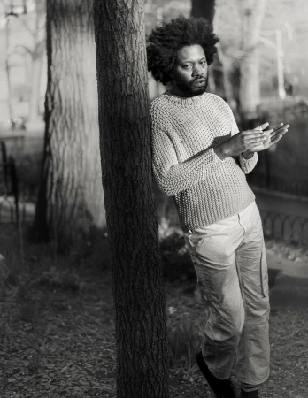 Black and white portrait of a Black adult male leaning against a tree trunk in an outdoor setting, wearing a sweater and pants.