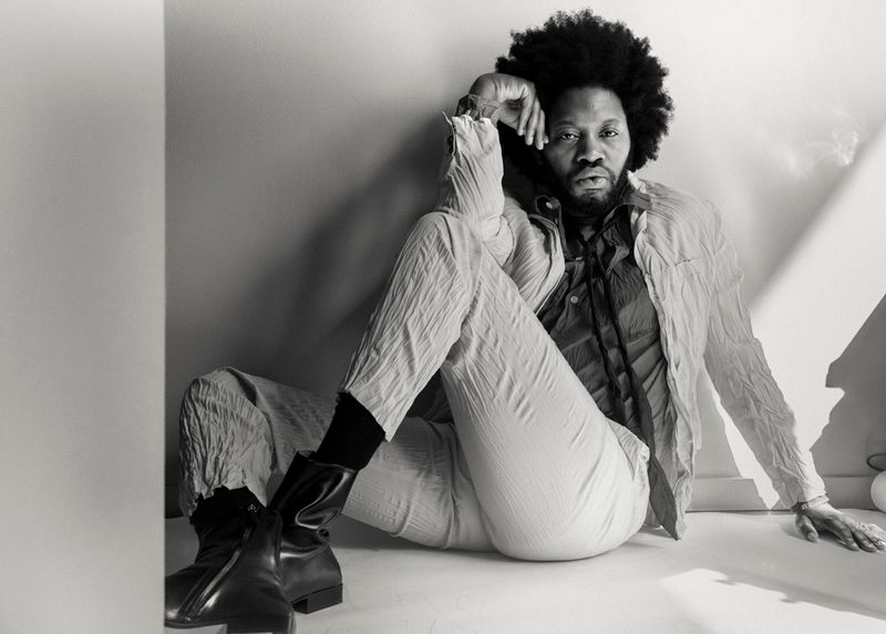Stylish portrait of a Black male model with afro hairstyle in wrinkled suit, boots, and black socks posing in a light-filled studio.