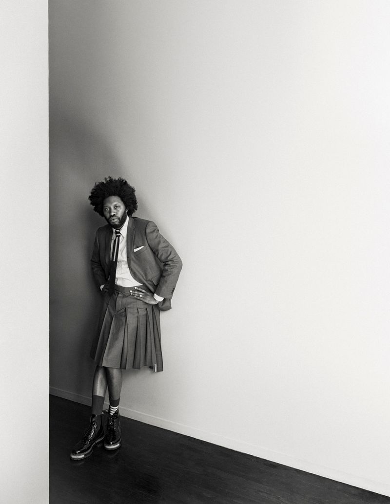Black man in a grey suit jacket and pleated kilt poses indoors against a grey and white wall in a fashion editorial.