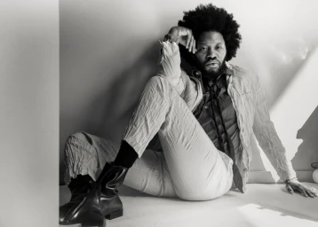 Black and white portrait of Jeremy O. Harris seated on the floor wearing a patterned shirt and light trousers.