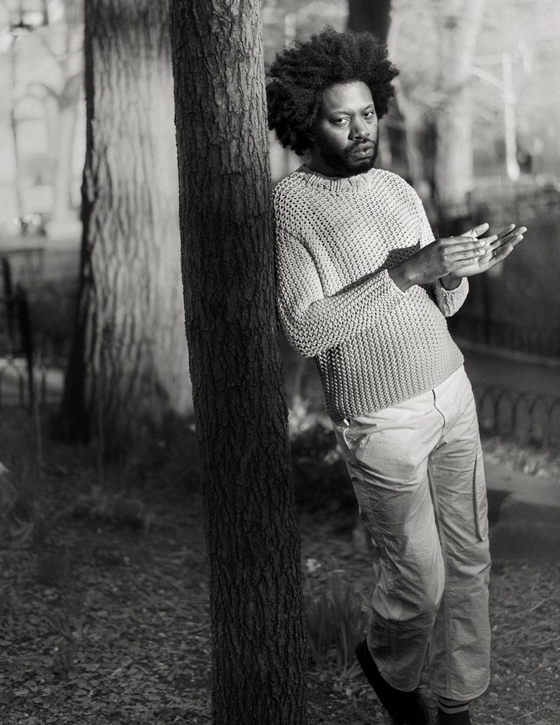 Black and white portrait of Jeremy O. Harris leaning against a tree trunk in an outdoor park setting.