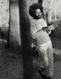 Black and white portrait of Jeremy O. Harris leaning against a tree trunk in an outdoor park setting.