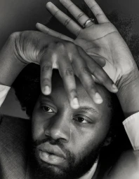 Close-up monochrome portrait of a man with his hands raised near his face, wearing a ring on one finger.