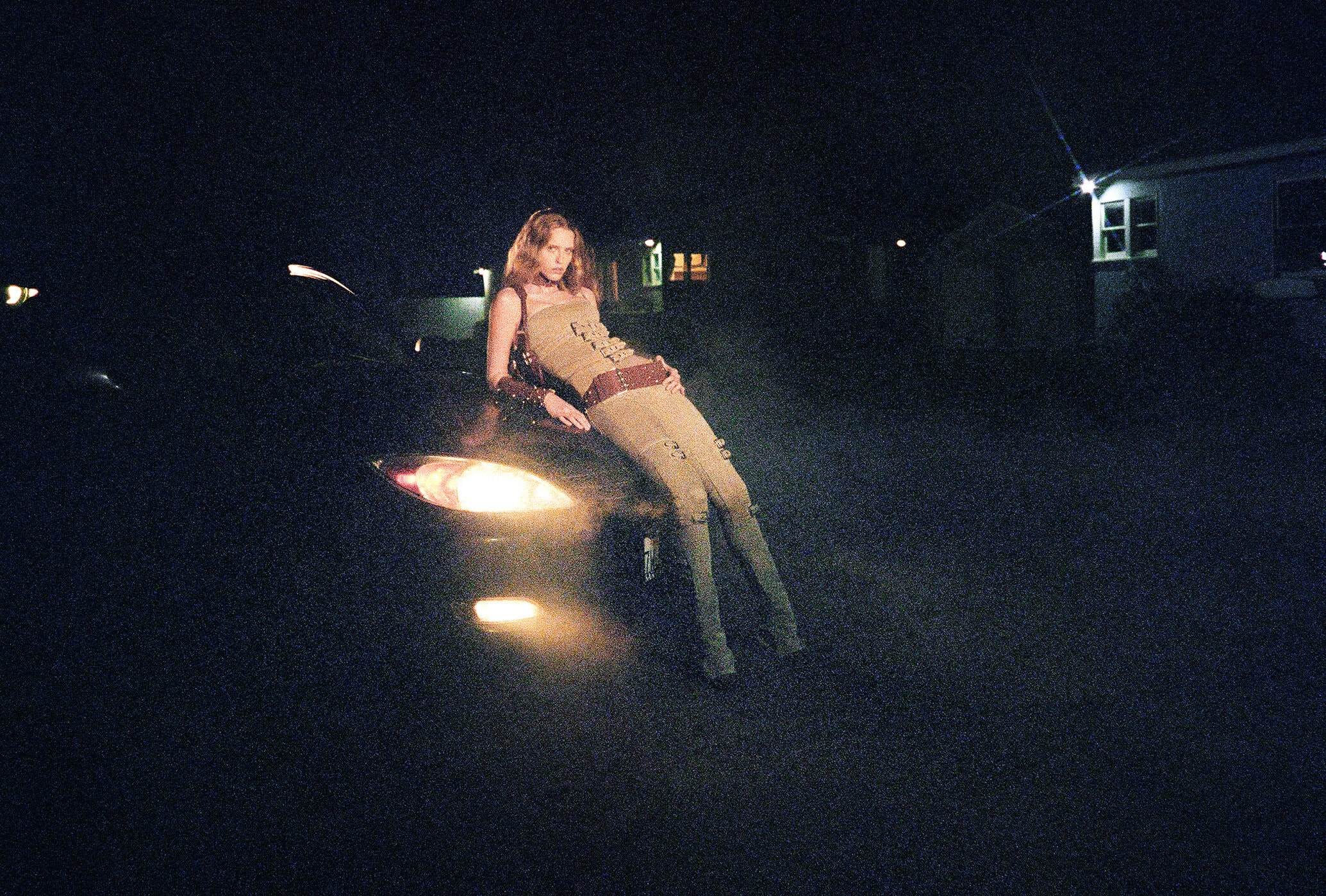 A woman in a tan outfit leans on the hood of a black car with its headlights on at night, standing outdoors against a dark background.