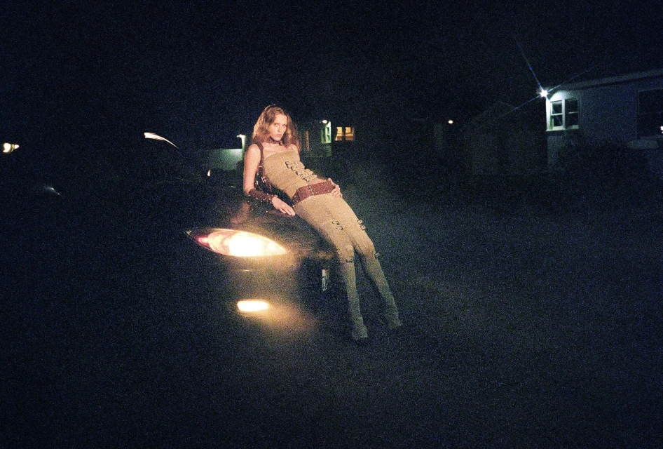 A woman in a tan outfit leans on the hood of a black car with its headlights on at night, standing outdoors against a dark background.