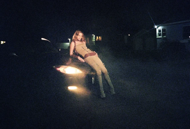 A woman in a tan outfit leans on the hood of a black car with its headlights on at night, standing outdoors against a dark background.