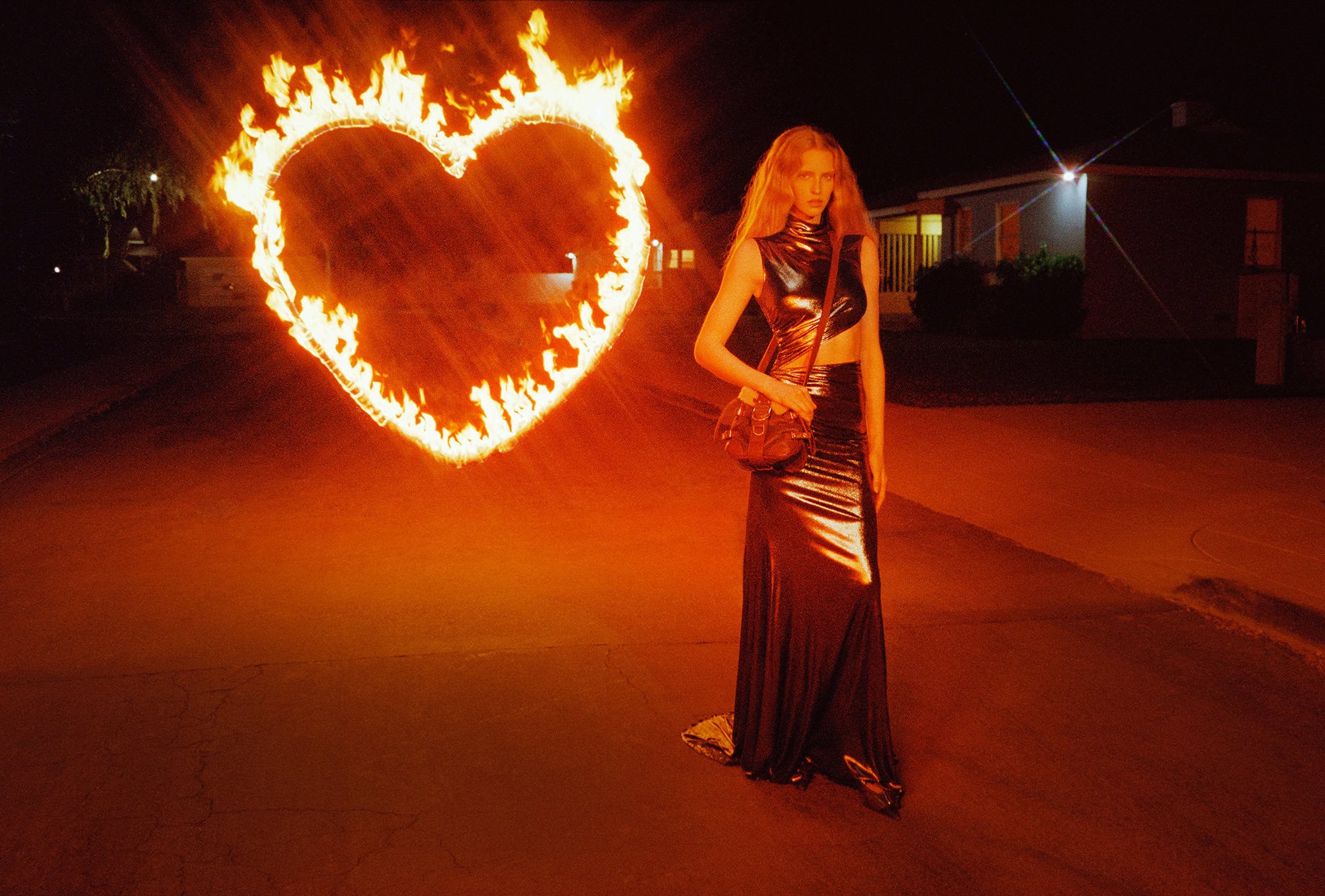 Woman poses in metallic dress next to a burning heart-shaped pyre on an empty street at night, illuminated by orange hues from the flames.