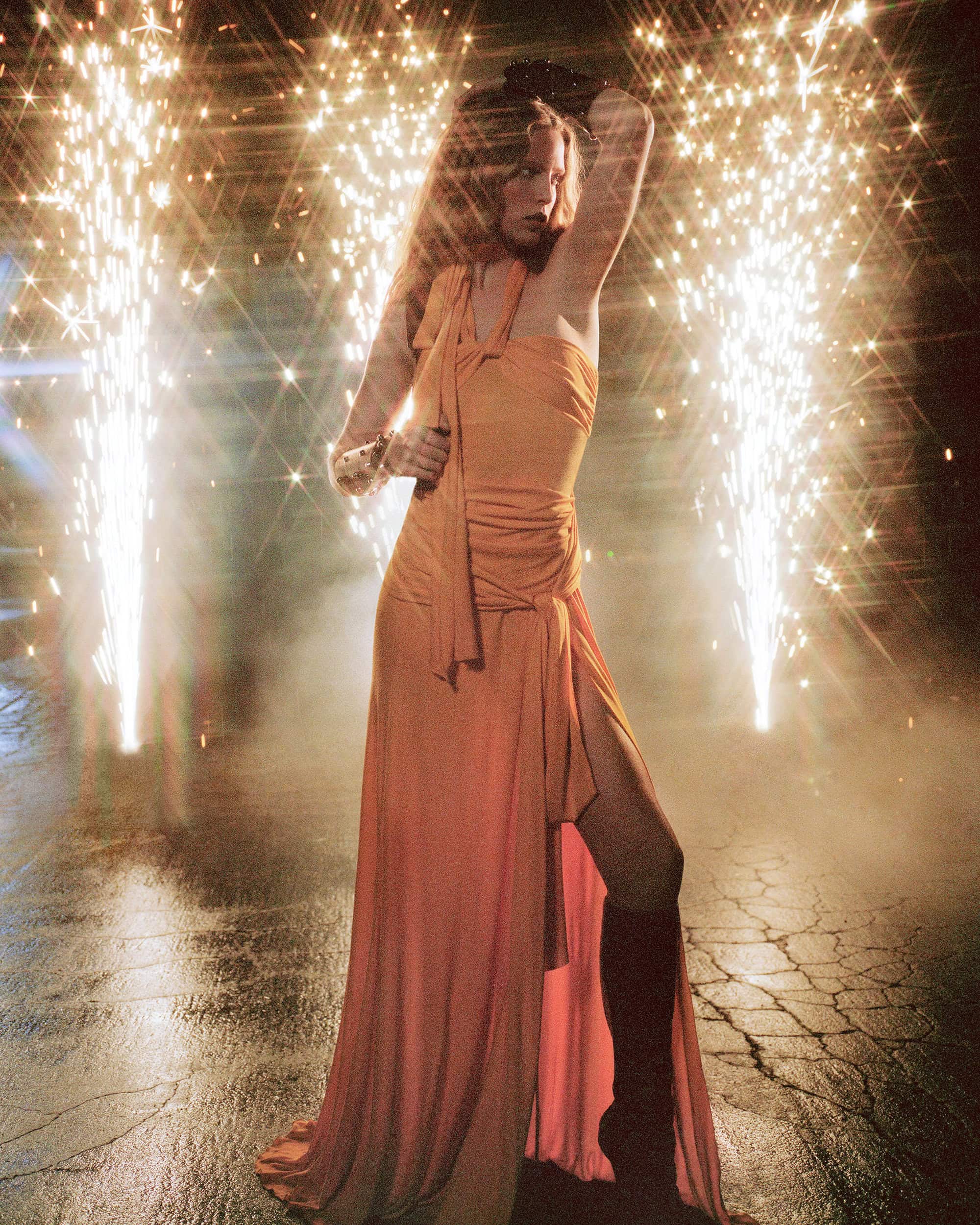 Woman in flowing coral gown poses in an outdoor setting with fireworks and smoke in the background creating a dramatic fashion image.