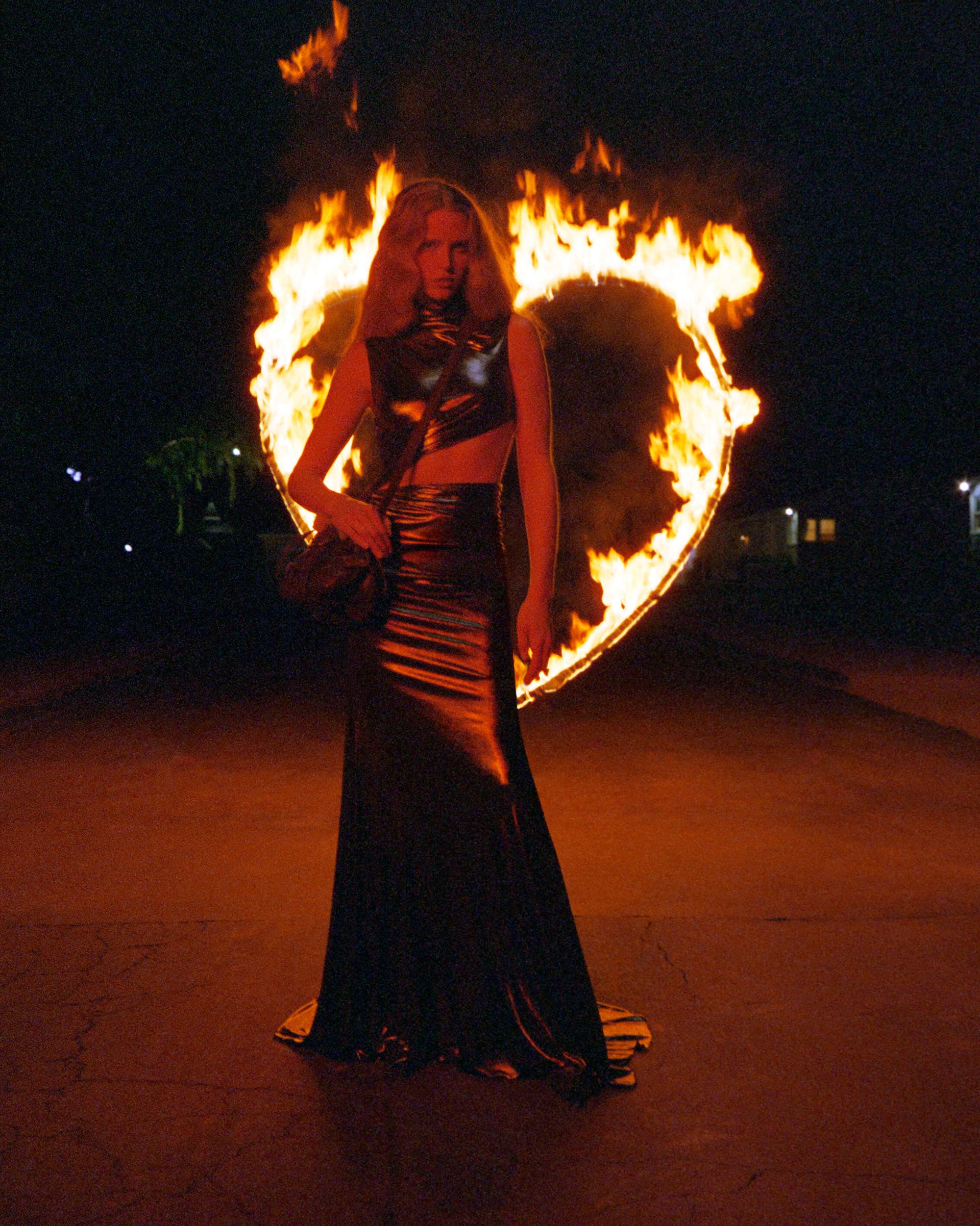 A woman in a black dress poses in front of a burning heart-shaped pyrotechnic display at night, with a dark background.