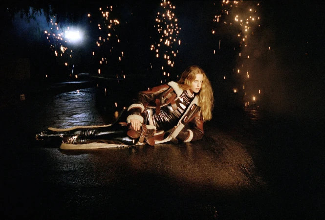 Model in futuristic jacket and leggings poses on wet ground surrounded by fireworks in a dark, outdoor setting at night.