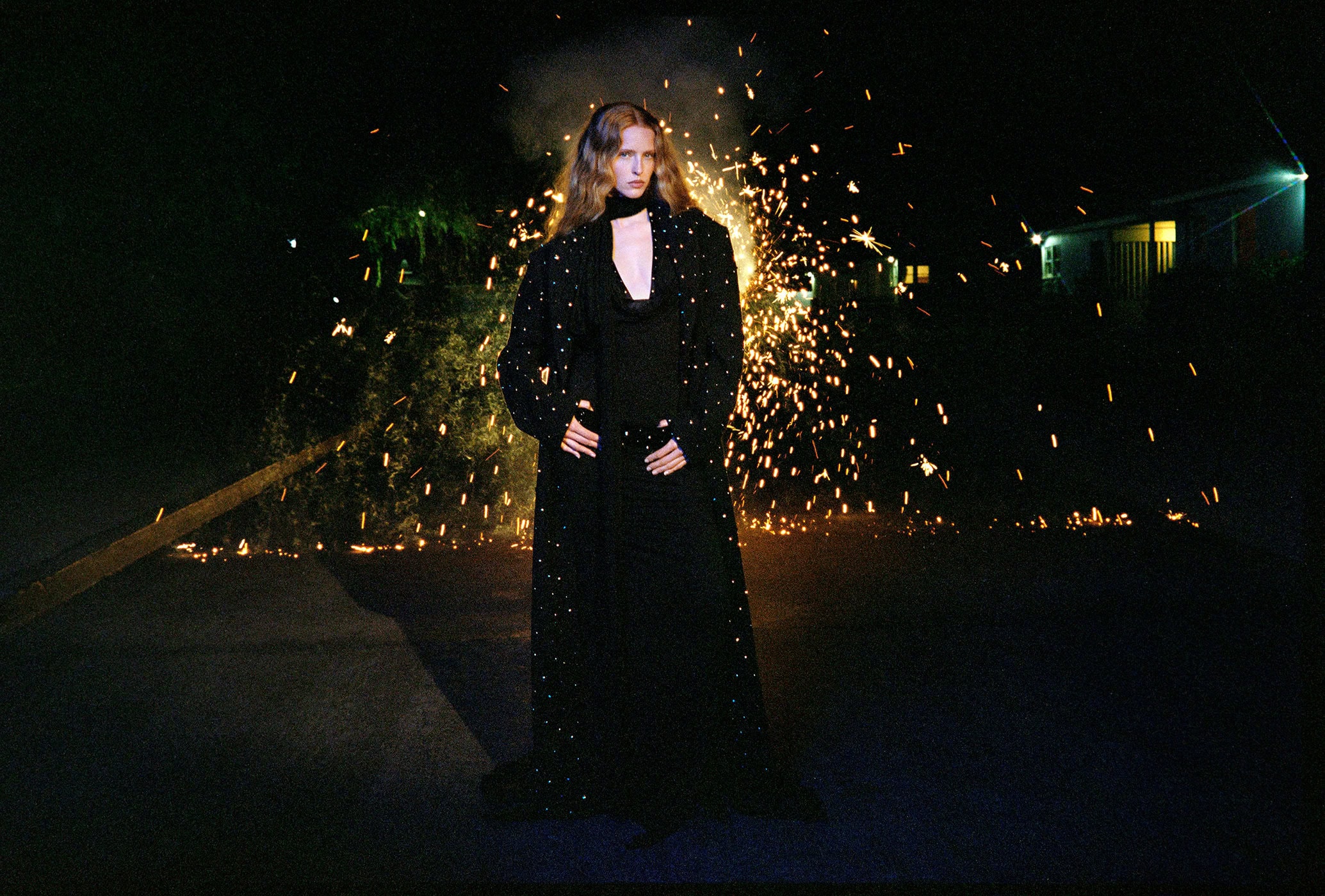 A woman in a long black coat stands on an asphalt road at night, illuminated by sparklers, with a building in the background.