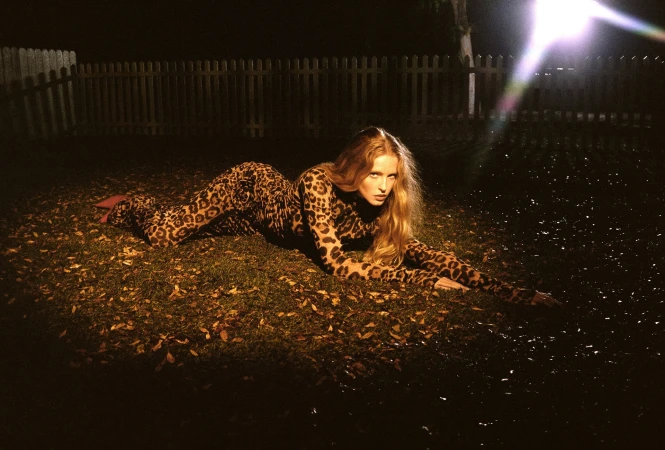 Model Abby Champion wearing a leopard print catsuit lying on a grassy lawn at night with a bright light flare.