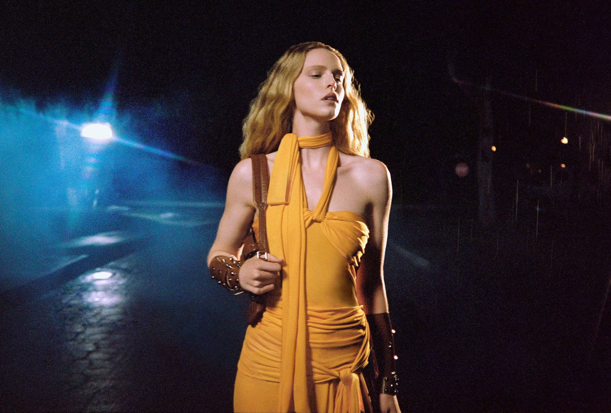 A woman in a yellow dress stands on an asphalt surface at night, wearing a shoulder bag, lit by a distant street light.