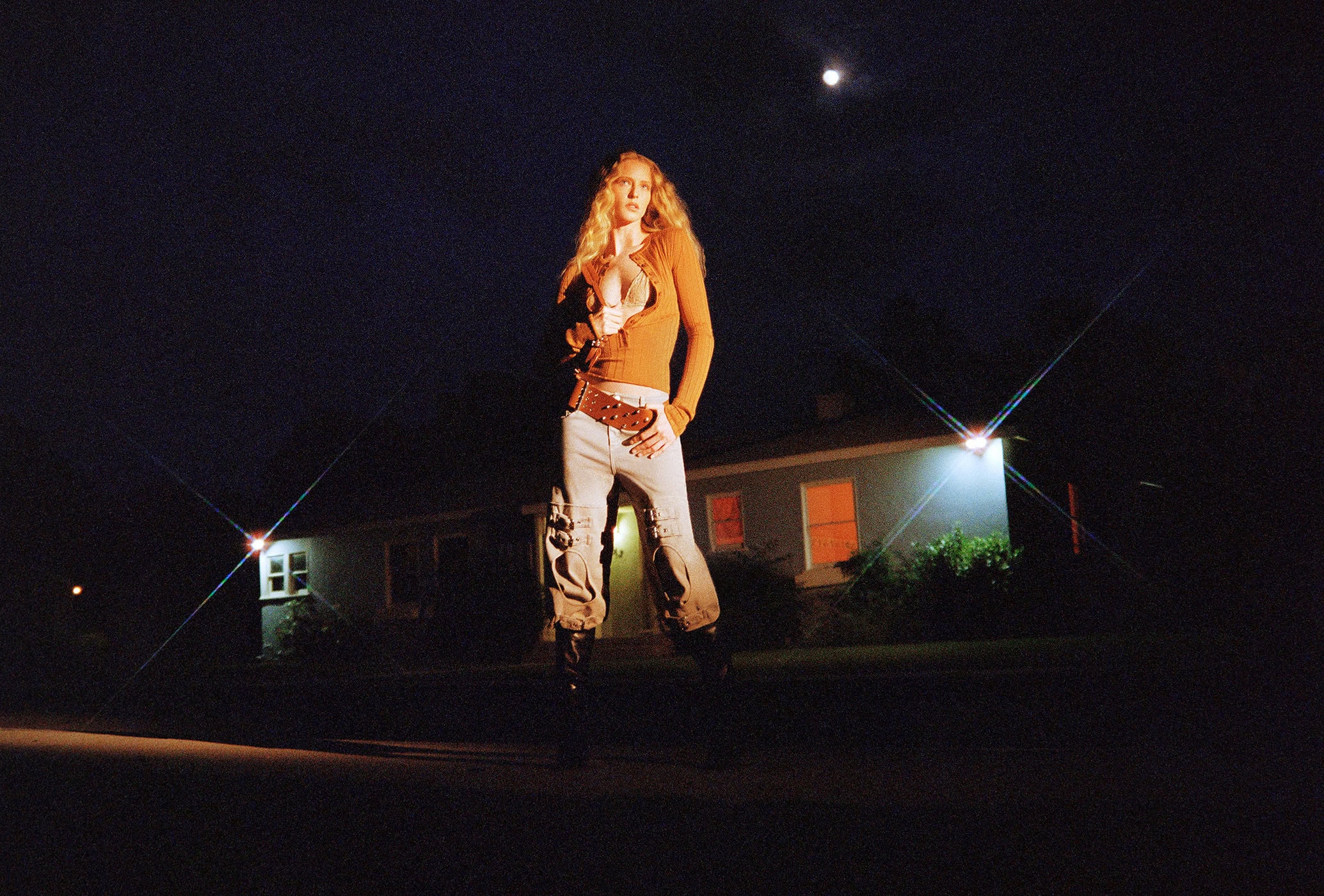 A blonde woman poses in an unbuttoned shirt and jeans in front of a house at night under the moon, illuminated by harsh outdoor lights.