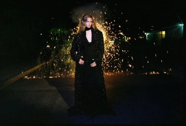 Model Abby Champion wearing an embellished long black coat standing outdoors at night with sparks erupting behind her.