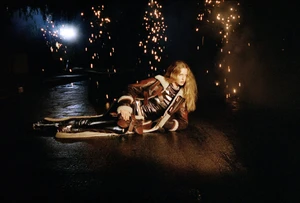 Model lying on an outdoor dark path wearing a shearling-trimmed leather jacket with scattered warm sparks around her.