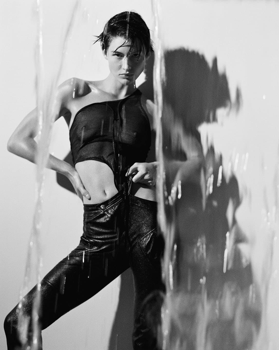 Monochrome portrait of a woman in wet hair and sheer top, standing in front of a water curtain with hands on hips, looking at the camera.
