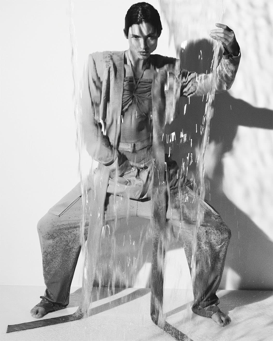 Monochrome fashion shot of a man in a coordinated outfit standing underneath a stream of flowing water in a bright studio.