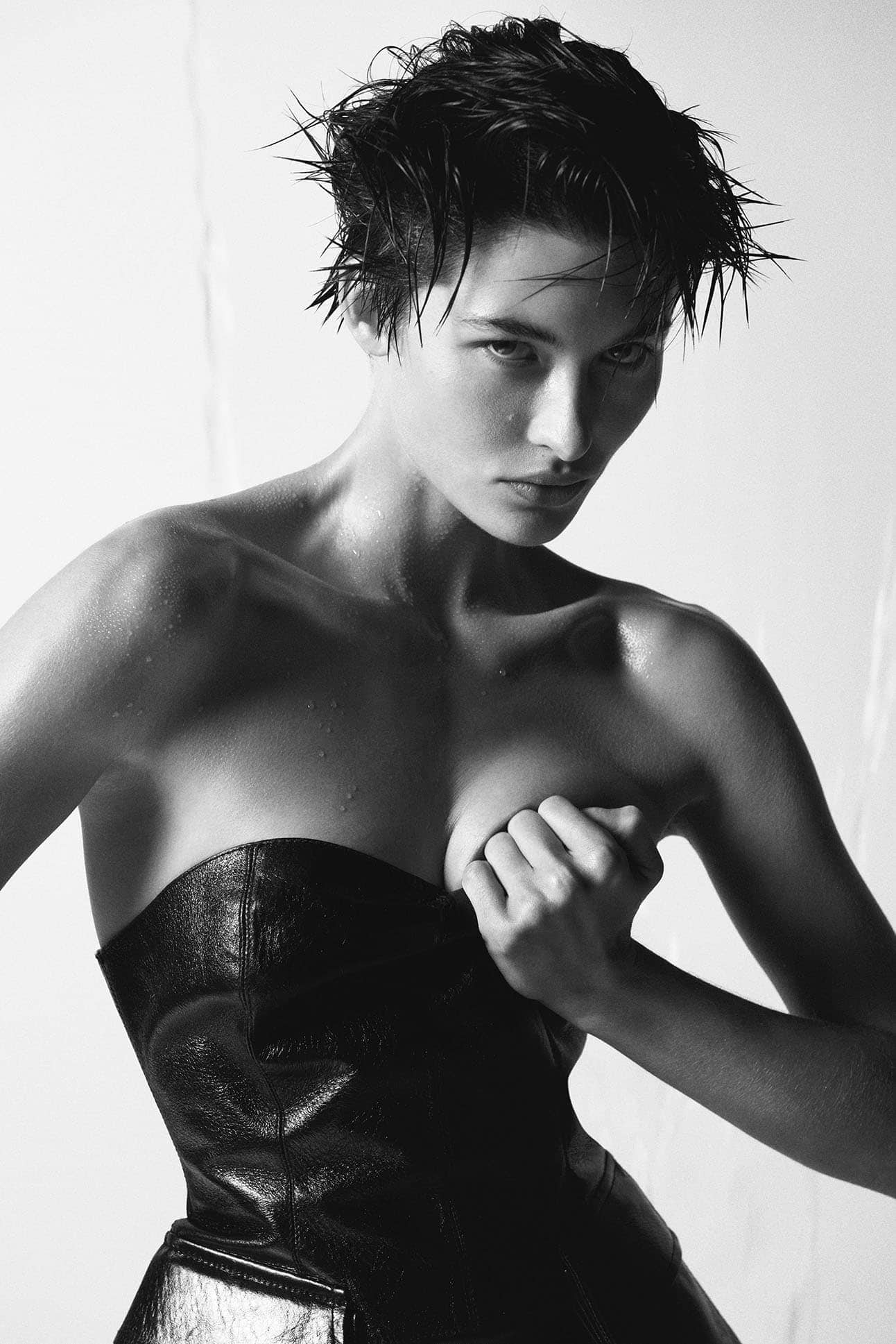 Monochrome fashion portrait of a woman with a wet pixie haircut, wearing a black strapless top, posing in front of a light backdrop.