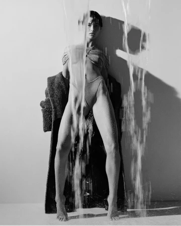 Black and white fashion shot of a female model under falling water, wearing a bodysuit and standing beside a coat.