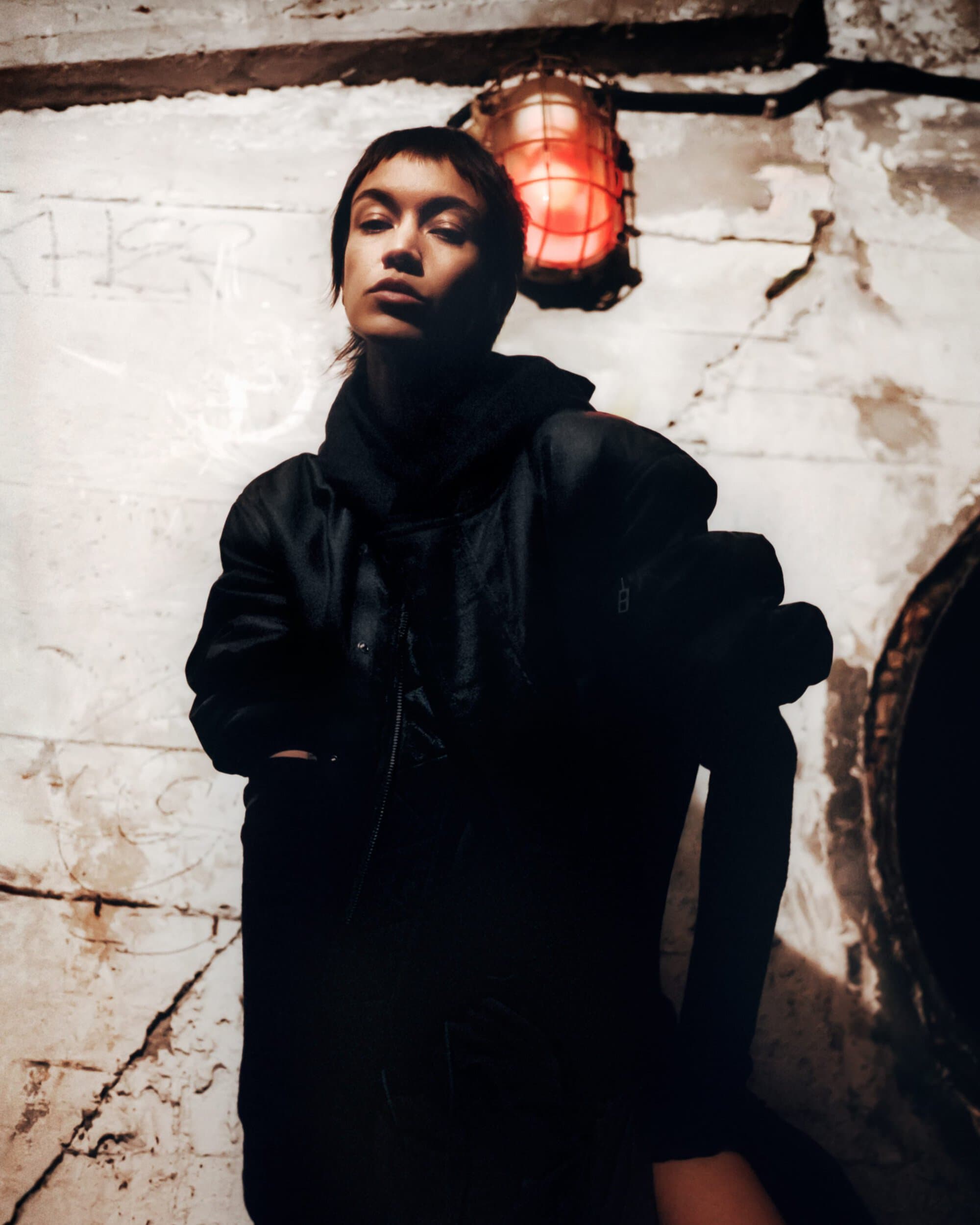 Young woman with short brown hair poses indoors near a bright red light, wearing a dark hoodie and jacket against a textured concrete wall.