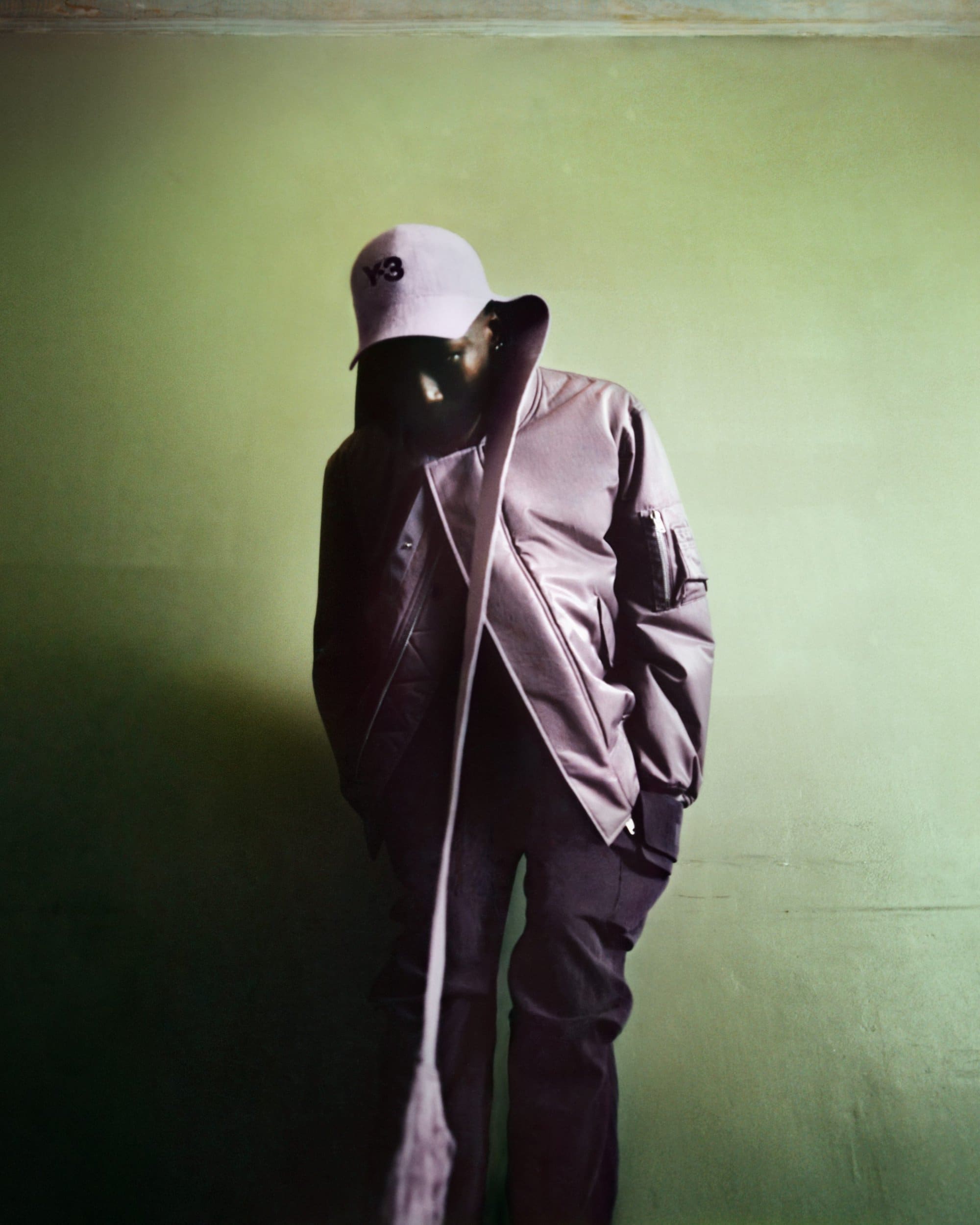 A person poses indoors against a green wall, wearing a black bomber jacket, dark pants, and a baseball cap with a Y-3 logo.